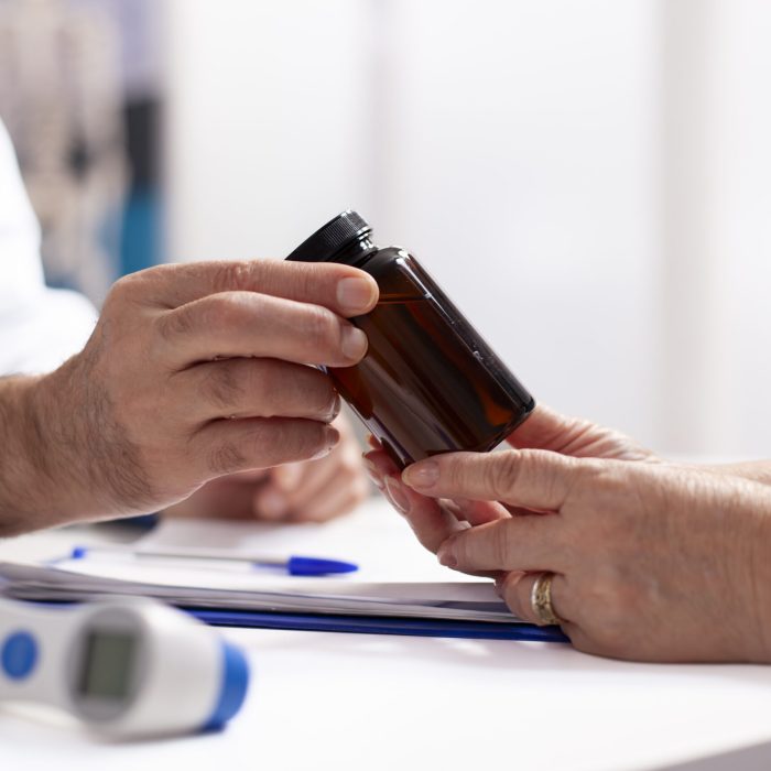 Closeup of white person in lab coat giving medicine to pensioner patient after medical exams. Selective focus of caucasian people holding pill bottle on table with thermometer and clipboard nearby.